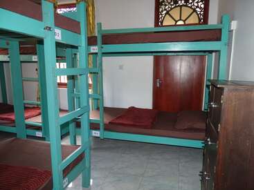 A imagem mostra um quarto simples de albergue com beliches azul-petróleo, roupa de cama marrom, uma cômoda de madeira, piso de ladrilho, uma porta de madeira e janelas com cortinas amarelas.