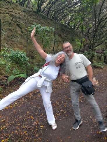 Uma mulher alegre de branco posa divertidamente com um homem sorridente de cinza, ambos em pé em uma trilha arborizada cercada por vegetação exuberante e árvores altas.