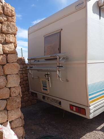 A white camper van is parked near a stone wall under a partly cloudy sky. The vehicle has a rear window, bike rack, and visible license plate.