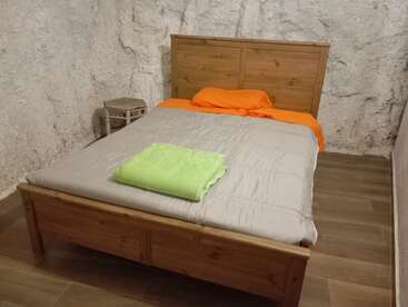 A cozy bedroom with a wooden bed, orange bedding, and a green towel. The room features textured white stone walls and a small rustic side table.