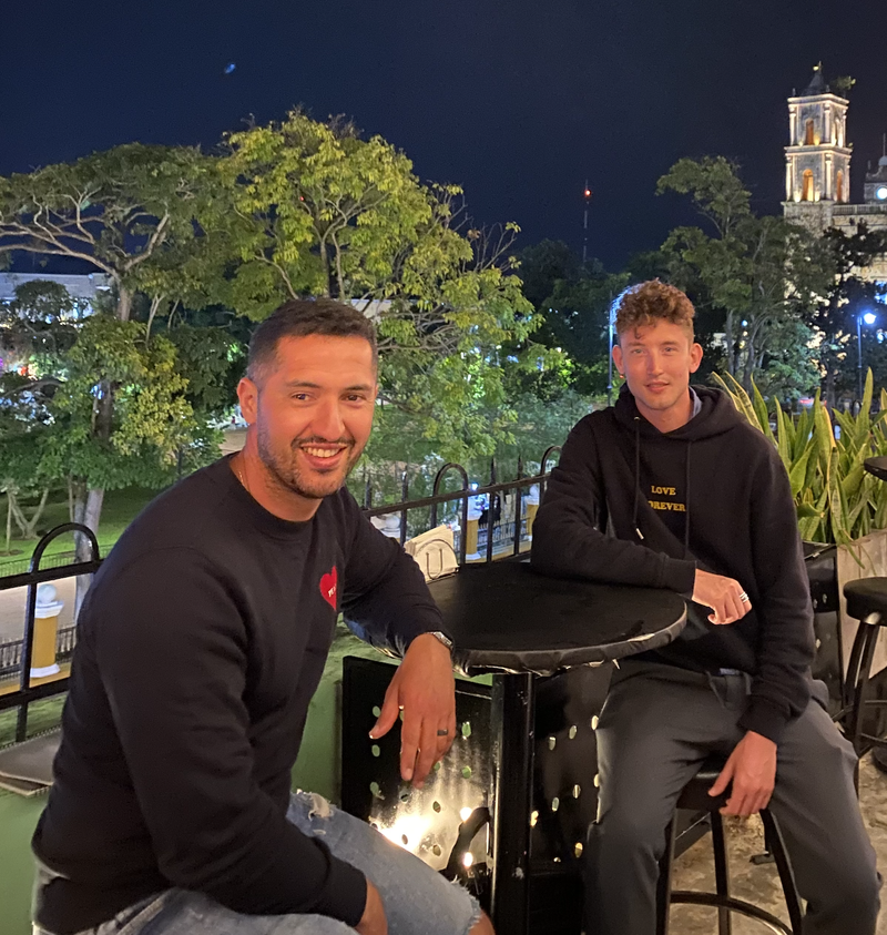 Dois caras estão sentados à noite numa mesa redonda ao ar livre, sorrindo. Árvores e um prédio iluminado aparecem ao fundo, criando uma vibe tranquila.