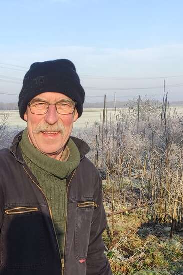 Un hombre con gafas, gorro negro y jersey verde está de pie al aire libre en un campo helado en un soleado día de invierno, sonriendo a la cámara.