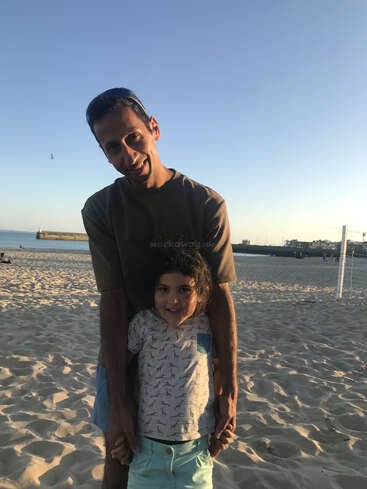 Un homme et un enfant se tiennent joyeusement sur une plage de sable au coucher du soleil. L'homme se penche en avant, souriant, tout en tenant l'enfant affectueusement par derrière.