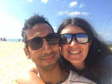 Un couple heureux portant des lunettes de soleil sourit en posant pour un selfie sur une plage ensoleillée. Le ciel bleu et le sable créent une atmosphère lumineuse et joyeuse.