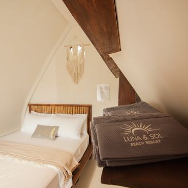 Um quarto aconchegante e rústico apresenta uma cama bem arrumada com lençóis brancos, uma cabeceira de tecido, travesseiros decorativos, iluminação suave e toalhas da marca Luna & Sol Beach Resort.