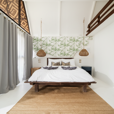 Um quarto aconchegante com tema tropical apresenta uma cama de madeira, roupa de cama branca, luminárias pendentes de tecido, decoração de parede com folhas, cortinas cinzas e um tapete de fibra natural sobre piso branco.