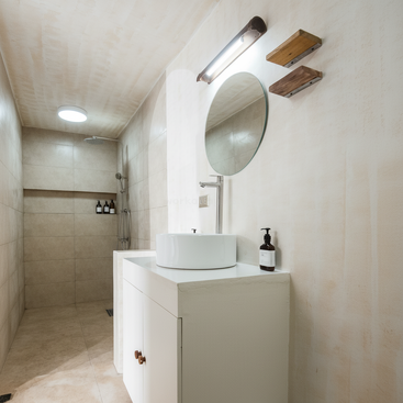 Um banheiro minimalista com espelho redondo, pia moderna, prateleiras na parede, área de banho em azulejo bege com chuveiro tipo ducha e iluminação sutil para um visual limpo.