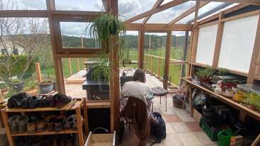 Una persona se sienta en el interior de un luminoso invernadero de madera lleno de macetas, zapatos y material de jardinería, con vistas a un exuberante paisaje verde a través de grandes ventanales.