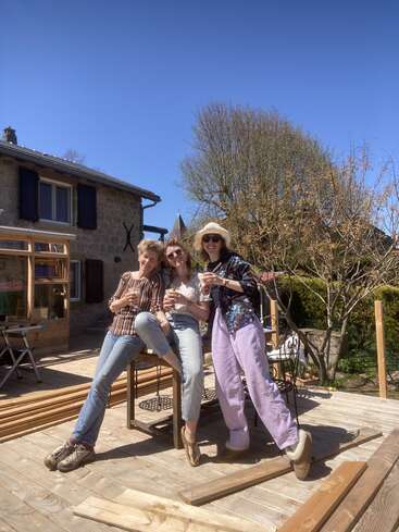 Tres mujeres disfrutan de unas copas al aire libre en un día soleado, sonrientes y relajadas en una terraza de madera cerca de una casa rústica de piedra, rodeadas de árboles y materiales de construcción.