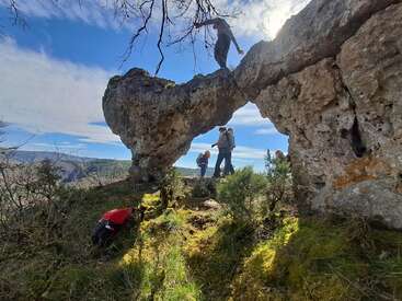 A group of hikers explores a rocky natural arch in a scenic landscape under a bright blue sky, surrounded by mossy ground, bushes, and distant mountains.