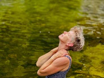 A woman stands in water with her head tilted back, eyes closed, hands on neck, appearing peaceful and content. Green water provides a natural backdrop.