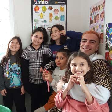 The image depicts a group of children and an adult posing for a photo in a classroom, with a poster of clothing items in the background.