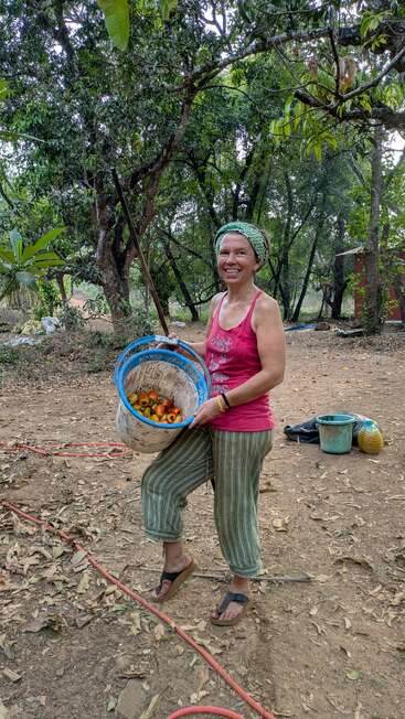Une femme souriante se tient debout à l'extérieur, dans une zone boisée, vêtue de vêtements décontractés et d'un foulard, tenant un seau rempli de fruits cueillis, profitant de la nature.
