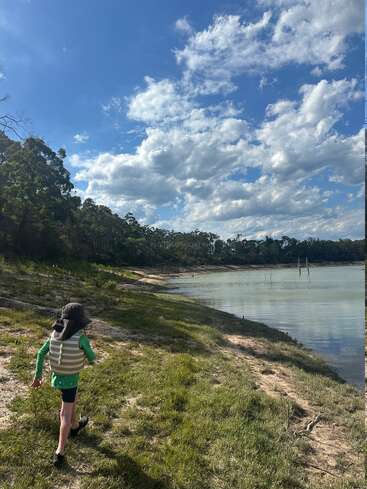 Un niño con sombrero y chaleco camina por la orilla cubierta de hierba de un lago, bajo un cielo azul brillante salpicado de nubes, rodeado de árboles y naturaleza.