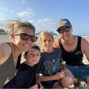 Eine lächelnde vierköpfige Familie genießt gemeinsam ein Eis an einem sonnigen Strand, gekleidet in lässige Sommerkleidung, mit blauem Himmel und Sand im Hintergrund.