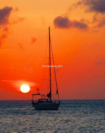 Das Bild zeigt eine ruhige Szene mit einem Segelboot, das bei Sonnenuntergang auf dem Meer treibt, während die Sonne dahinter untergeht. Der Himmel ist orange und bewölkt.