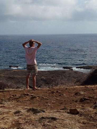 Das Bild zeigt einen Mann, der an einem felsigen Strand steht und mit erhobenen Händen auf den Ozean hinausblickt. Er trägt ein rosa Hemd und karierte Shorts.