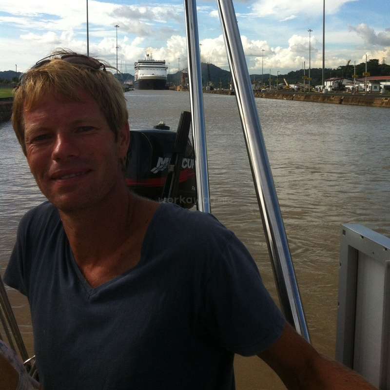 Das Bild zeigt einen Mann, der auf einem Boot steht, mit einem Kanal und einem Kreuzfahrtschiff im Hintergrund. Der Mann trägt ein blaues Hemd.