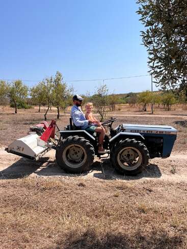 A man and a young child are sitting together on a blue tractor in a dry, rural landscape with scattered trees under a clear blue sky.