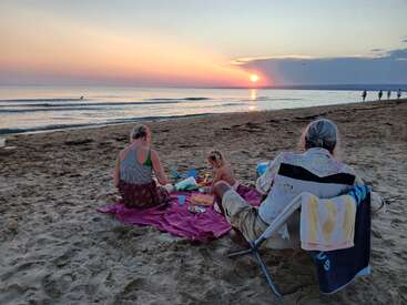 Three people enjoy a relaxing picnic on a sandy beach at sunset. Soft waves, warm light, and a tranquil atmosphere create a beautiful, peaceful summer evening.