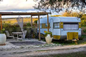 A cozy vintage caravan sits beside an outdoor seating area with chairs and a table under a pergola. Potted plants, bicycle, and countryside landscape complete the scene.