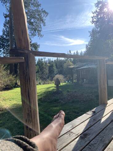 Eine Person entspannt sich barfuß auf einer Holzveranda und genießt einen sonnigen Tag. Umgeben von Bäumen, einem Rasen, einem rustikalen Gebäude, friedlicher Natur und blauem Himmel über ihr.