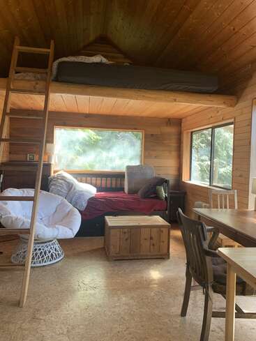 Diese gemütliche Holzhütte verfügt über ein Hochbett, das über eine Leiter erreichbar ist, ein Tagesbett, große Fenster, einen bequemen Sessel, eine Holzkiste und eine kleine Essecke.