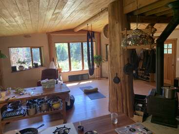 Dieser gemütliche, rustikale Wohnraum besticht durch Holzbalken, große Fenster mit Blick ins Grüne, Küchenutensilien, einen Holzofen, hängende Pfannen und überall farbenfrohe Zimmerpflanzen.