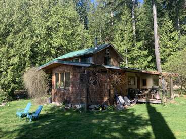 Eine gemütliche Holzhütte steht in einer üppigen, bewaldeten Gegend. Zwei blaue Stühle stehen auf grünem Gras. Sonnenlicht fällt durch die hohen Bäume, die das Haus umgeben.