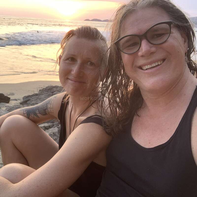 Zwei lächelnde Menschen sitzen bei Sonnenuntergang dicht beieinander an einem felsigen Strand. Die Meereswellen und das goldene Sonnenlicht schaffen eine friedliche, warme und fröhliche Atmosphäre.