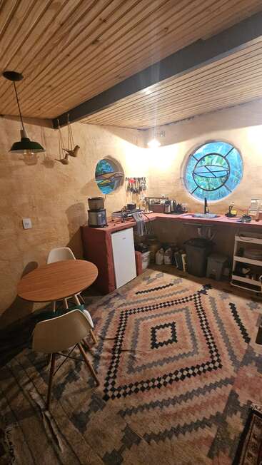 Esta acogedora cocina cuenta con ventanas redondas, techo de madera, alfombra estampada, electrodomésticos compactos, una pequeña mesa de comedor, tazas colgantes, paredes de tonos tierra y un ambiente rústico y artesanal en todo el espacio.