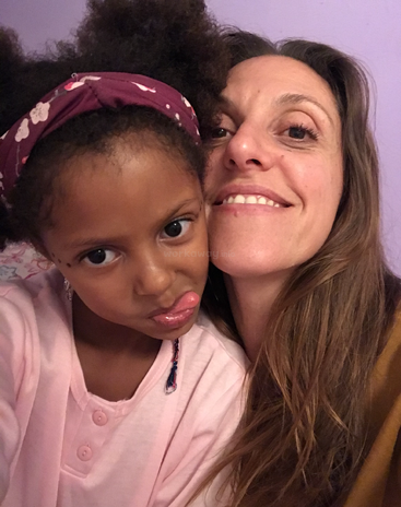 Una niña y una mujer posan juntas, sonriendo y haciendo muecas graciosas. La niña lleva una diadema y una camiseta rosas, y el fondo es morado.