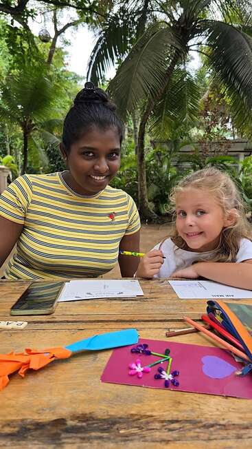 Two smiling girls sit at a wooden table outdoors, crafting and writing. Surrounded by lush greenery, art supplies and paper flowers decorate the table, creating a cheerful scene.