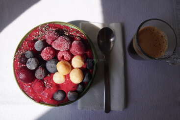 A vibrant smoothie bowl topped with frozen berries, macadamia nuts, and chia seeds, alongside an espresso and a spoon on a napkin. Bright, inviting breakfast.