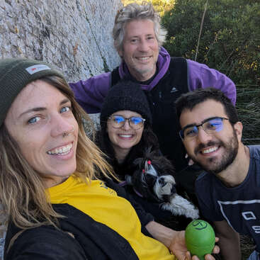 The image shows four people and a dog posing for a selfie in front of a rock wall, with a grassy area and bushes in the background.