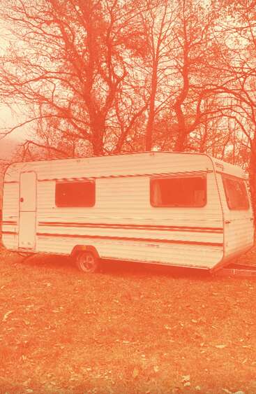Uma caravana vintage branca fica em um terreno gramado, cercada por árvores sem folhas. Toda a cena é lavada com um filtro laranja-avermelhado, criando uma atmosfera nostálgica.