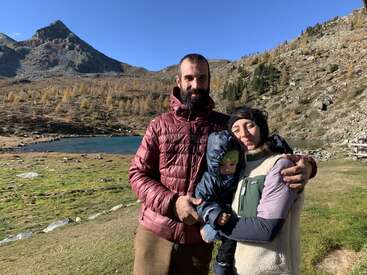 Uma família de três pessoas fica ao ar livre perto de um lago na montanha, agasalhada com roupas quentes, aproveitando o sol e a bela paisagem natural em um dia claro.