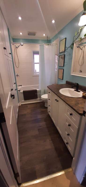 Das ist ein gemütliches, helles Badezimmer mit Holzboden, blauen Wänden, weißen Schränken, einer braunen Arbeitsplatte, einer Badewanne, einer Dusche, einer Toilette, einem Spiegel und Wandbildern.