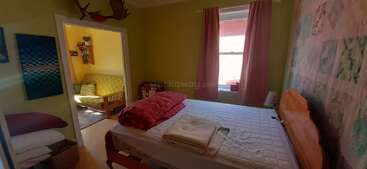 Ein gemütliches Schlafzimmer mit grünen Wänden, einem Bett mit Kissen und gefalteten Decken, einem Fenster mit rosa Vorhängen und einem angrenzenden Zimmer mit einer grünen Couch.