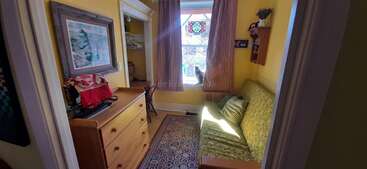 Dieser gemütliche, sonnendurchflutete Raum hat gelbe Wände, eine kleine Kommode, ein grünes Sofa, einen gemusterten Teppich, einen Schreibtisch, ein Fenster mit einer Katze, Kunstwerke und dekorative Regale mit Kleinigkeiten.