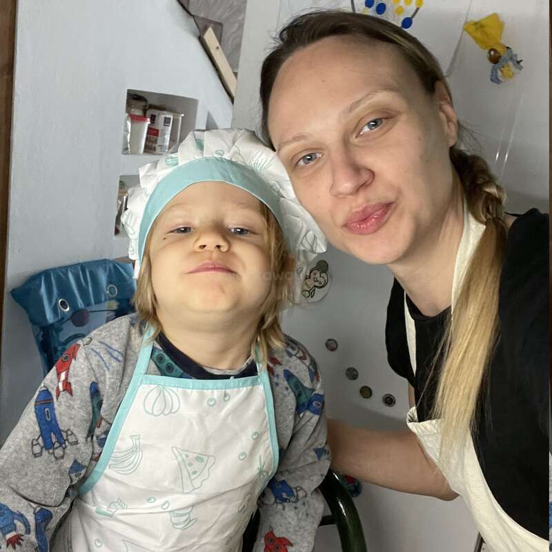 A imagem mostra uma mulher e uma criança em uma cozinha, ambas usando aventais e chapéus de chef, posando para uma foto em frente a uma geladeira branca com ímãs.