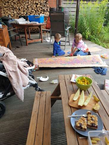 Two children paint on a large cardboard outdoors. Nearby, a table holds sliced melon, yellow plums, cake, and a stroller with a pink blanket. Cozy, creative atmosphere.