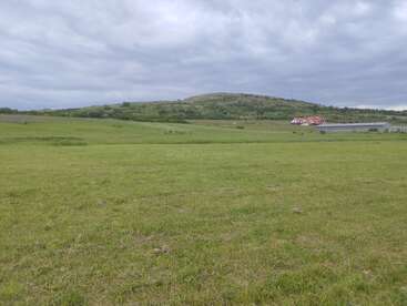A wide grassy field stretches toward a distant green hill. Two houses sit under a cloudy sky, surrounded by quiet countryside and open space. Peaceful landscape.
