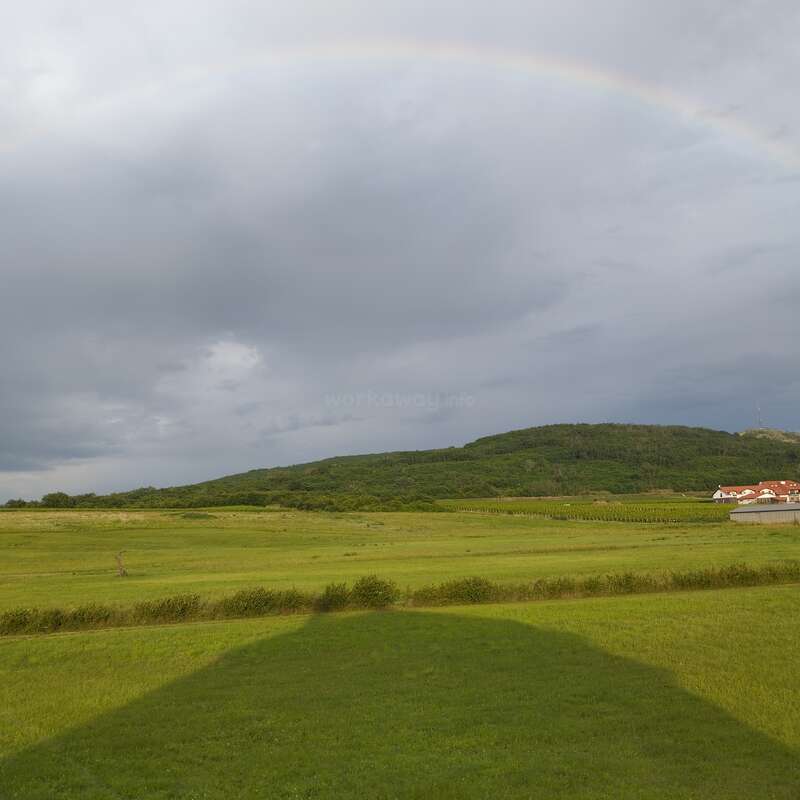 A faint rainbow arches over a lush green landscape, featuring a hill, cloudy sky, distant houses, and a large shadow stretching across the vivid grassy field.