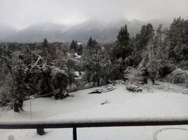 La imagen representa una serena escena invernal desde un balcón, con árboles cubiertos de nieve, una cordillera lejana y un ambiente tranquilo, con un sutil toque de niebla.
