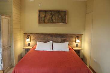 La imagen muestra un dormitorio con una cama, una cómoda y una mesilla de noche, y una gran ventana con vistas a una masa de agua y una cadena montañosa al fondo.