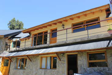 A two-story stone house with a corrugated metal roof and wooden accents, large windows, a balcony railing, and a clear blue sky in the background.