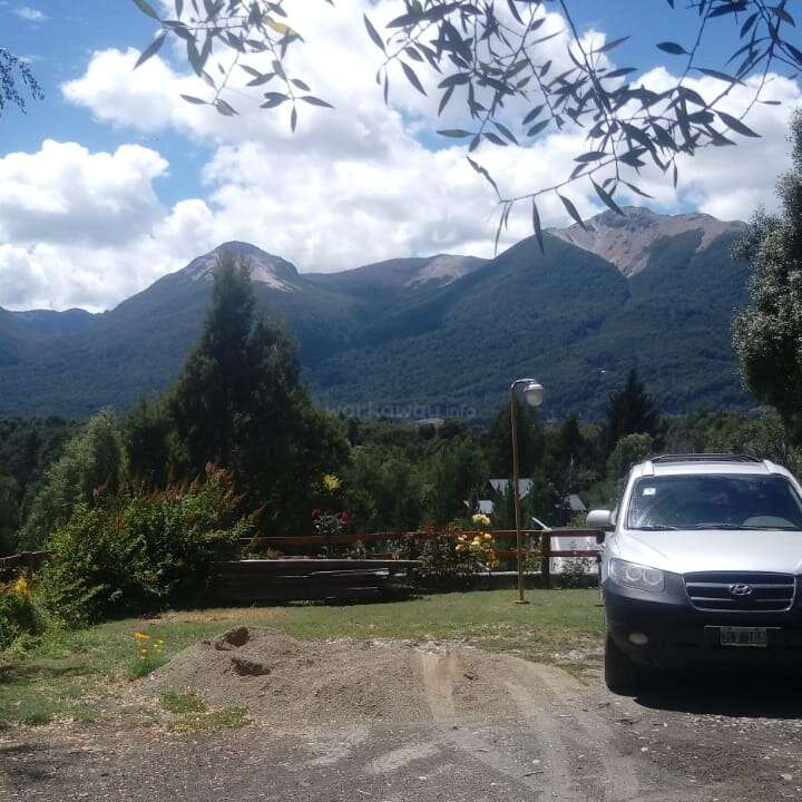 La imagen muestra un sereno paisaje montañoso con un todoterreno blanco aparcado en la entrada, rodeado de frondosos árboles y un pintoresco telón de fondo de colinas y montañas.