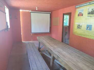 Dieses Bild zeigt ein einfaches Besprechungs- oder Klassenzimmer mit orangefarbenen Wänden, einem großen Holztisch und Bänken, einer Projektionsfläche und einem Bildungsposter an der Wand.