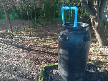 Ein großes schwarzes Fass mit blauen PVC-Rohren steht im Freien auf einem Betonsockel, umgeben von Bäumen und trockenem Laub, und sonnt sich im Morgenlicht.